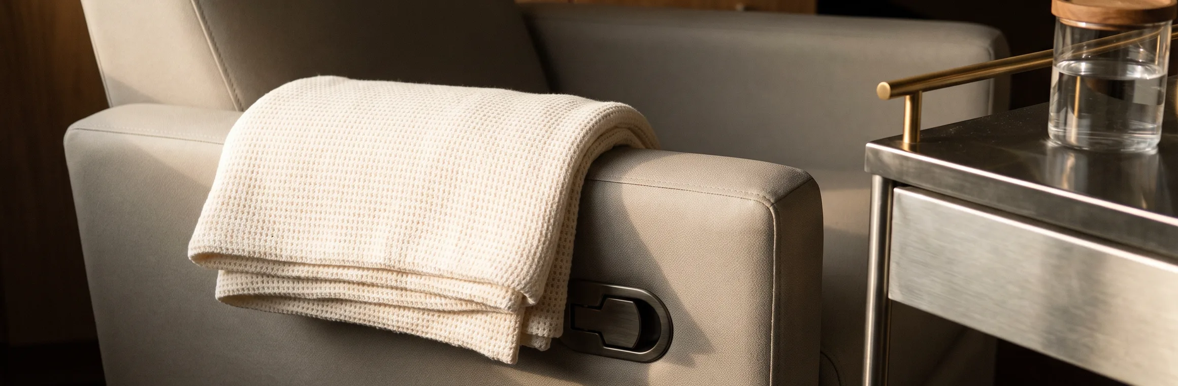 A folded cream cotton blanket over the arm of an upholstered recliner, with a stainless side trolley alongside carrying a covered glass of water.