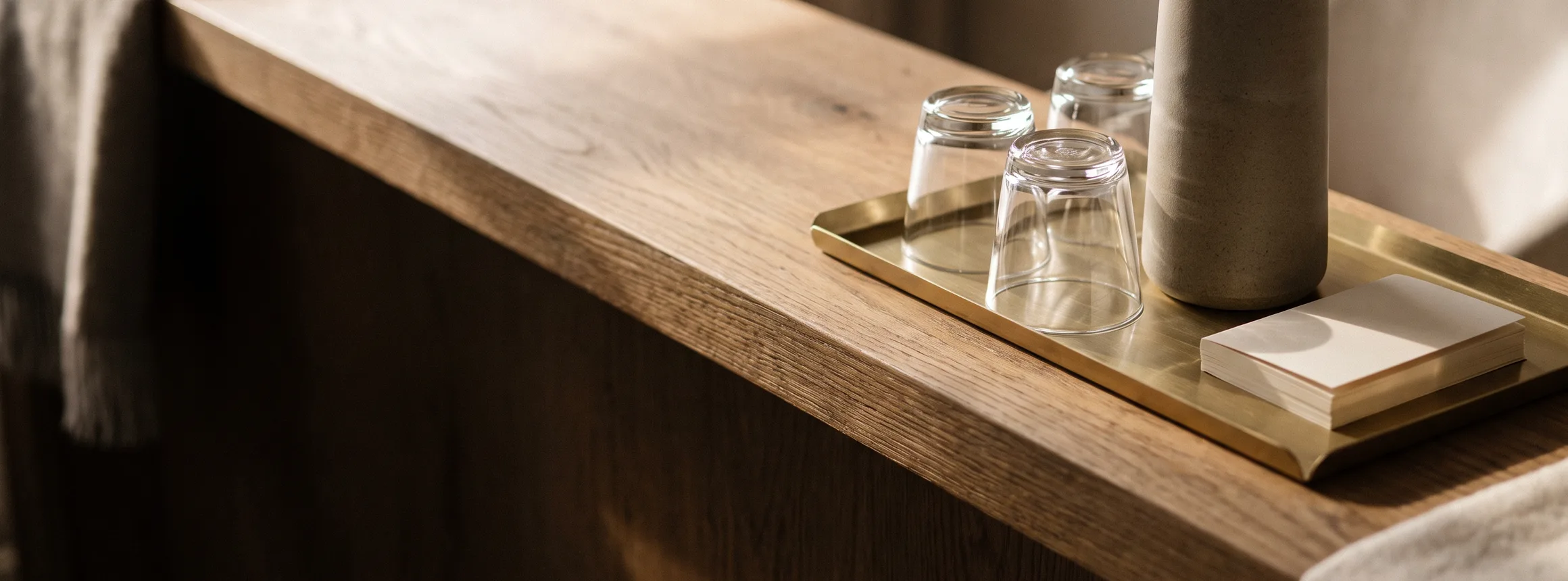 A stoneware carafe of water and two upturned glasses on a brass tray at the end of a warm oak counter, in morning light.
