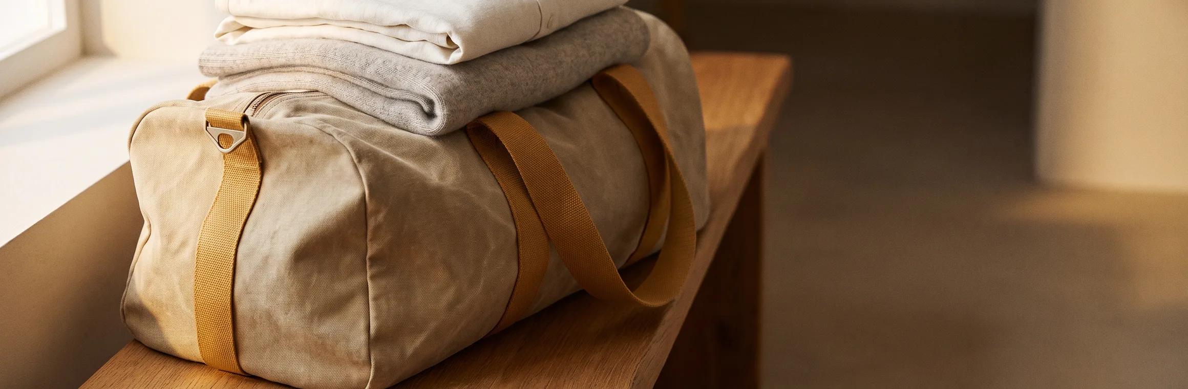 A packed canvas holdall with folded clothes resting on top, on a wooden bench in morning light, flat slip-on shoes on the floor beside it.